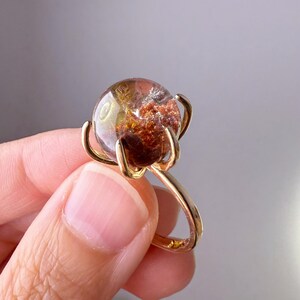 Könnte beinhalten: Ein goldfarbener Ring mit einem großen, runden Edelstein. Der Edelstein hat interne Einschlüsse in Braun-, Orange- und Klartönen. Er wird von vier gebogenen Zinken gehalten. Der Ring hat ein schlichtes, poliertes Design.
