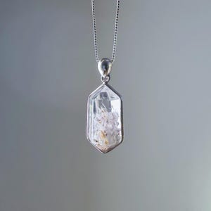 以下が含まれることがあります： シルバーカラーのベゼルとチェーンが付いた、透明なクリスタルペンダント。 クリスタルは六角形で、白と黄色のインクルージョンが含まれています。 チェーンは繊細なボールチェーンスタイルで、背景はソフトグレーです。