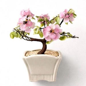 May include: A miniature bonsai tree with delicate pink blossoms and green leaves. The tree's brown trunk curves gracefully from a white square pot filled with small beige pebbles. The blossoms have a yellow center.