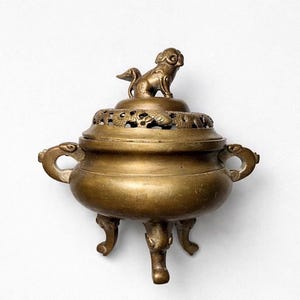 Puede incluir: Un quemador de incienso de bronce con una figura decorativa de perro en la tapa. El quemador tiene un cuerpo redondo con asas y se apoya en tres patas ornamentadas. La parte superior presenta intrincados diseños recortados.