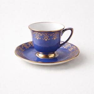 May include: A blue teacup and saucer set with gold filigree detailing. The teacup and saucer have a gold rim and a gold base. The teacup has a handle. The set is on a white background.