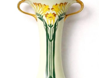 Op de afbeelding: Crèmekleurige vaas met geel bloemmotief en gouden accenten. De vaas heeft twee handvatten en een uitlopende voet. Het bloemmotief omvat gele bloemen met bruine centra en groene stelen. Een decoratief stuk voor de woondecoratie.