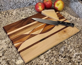 Op de afbeelding: Een houten snijplank met een golvend ontwerp gemaakt van verschillende soorten hout, waaronder walnoot, esdoorn en kers. Een mes en een stuk kaas liggen op het plank.