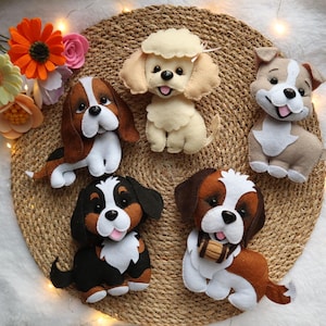 May include: Five handcrafted felt dog figurines in various breeds and colors, including a cream poodle, a brown and white hound, a tan puppy, a black and brown dog, and a brown and white dog with a small barrel, arranged on a round woven mat.