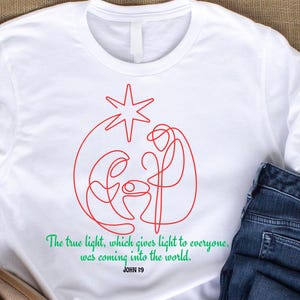 Puede incluir: Camiseta blanca con un dibujo lineal rojo de la escena de la Natividad y una estrella. El texto "The true light, which gives light to everyone, was coming into the world. JOHN 19" está impreso en verde. La camiseta se muestra con vaqueros azules.