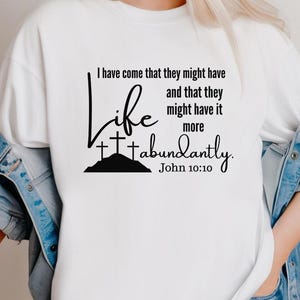 May include: White t-shirt featuring a black graphic. The design includes the word "Life", three crosses on a hill, and the text "I have come that they might have...John 10:10."