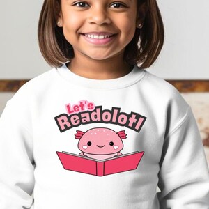 May include: White sweatshirt with a pink axolotl reading a book. The text on the shirt says "Let's Readolottl!"