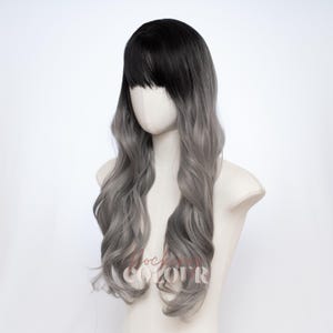 May include: A long wavy wig featuring a dark black root color that transitions into a light silver gray shade. The wig includes straight bangs that frame the forehead and long, loose curls that cascade down the sides and back of the head.