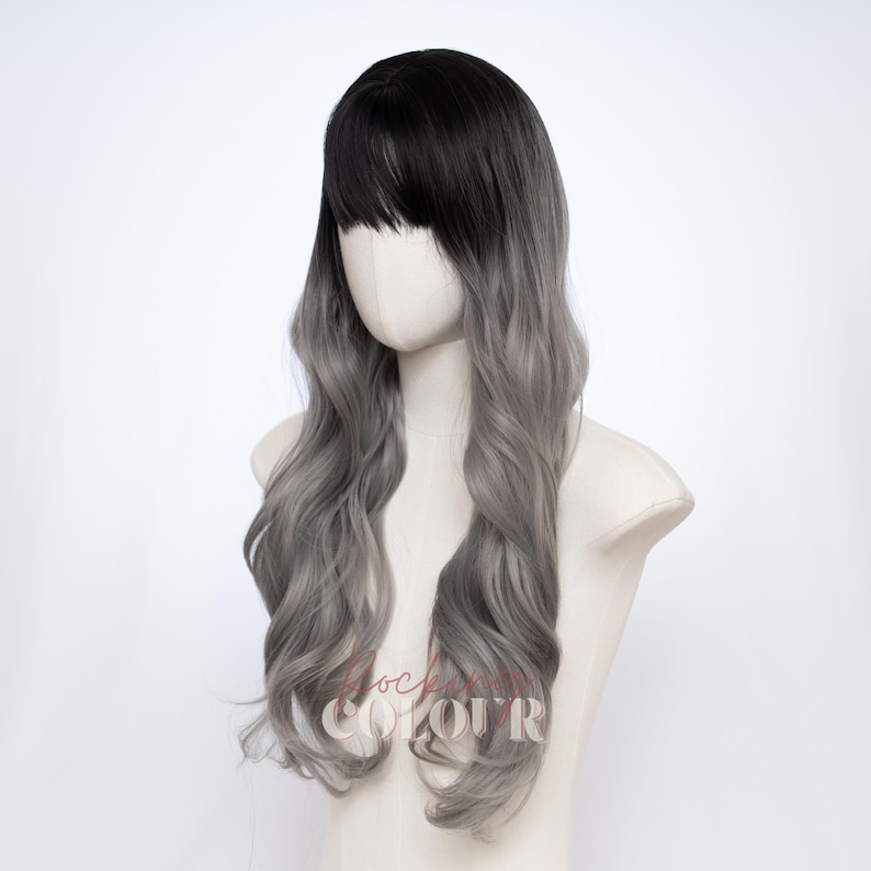 May include: A long wavy wig featuring a dark black root color that transitions into a light silver gray shade. The wig includes straight bangs that frame the forehead and long, loose curls that cascade down the sides and back of the head.
