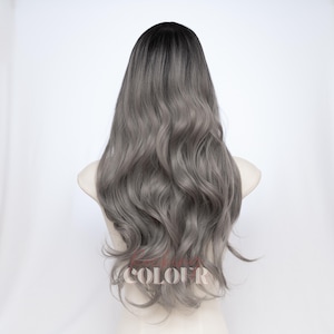 May include: A long, wavy wig featuring a dark charcoal root that transitions into a lighter ash gray tone. The hair has a soft, flowing texture with loose curls cascading down the back. The text COLOUR is visible in a light font at the bottom.