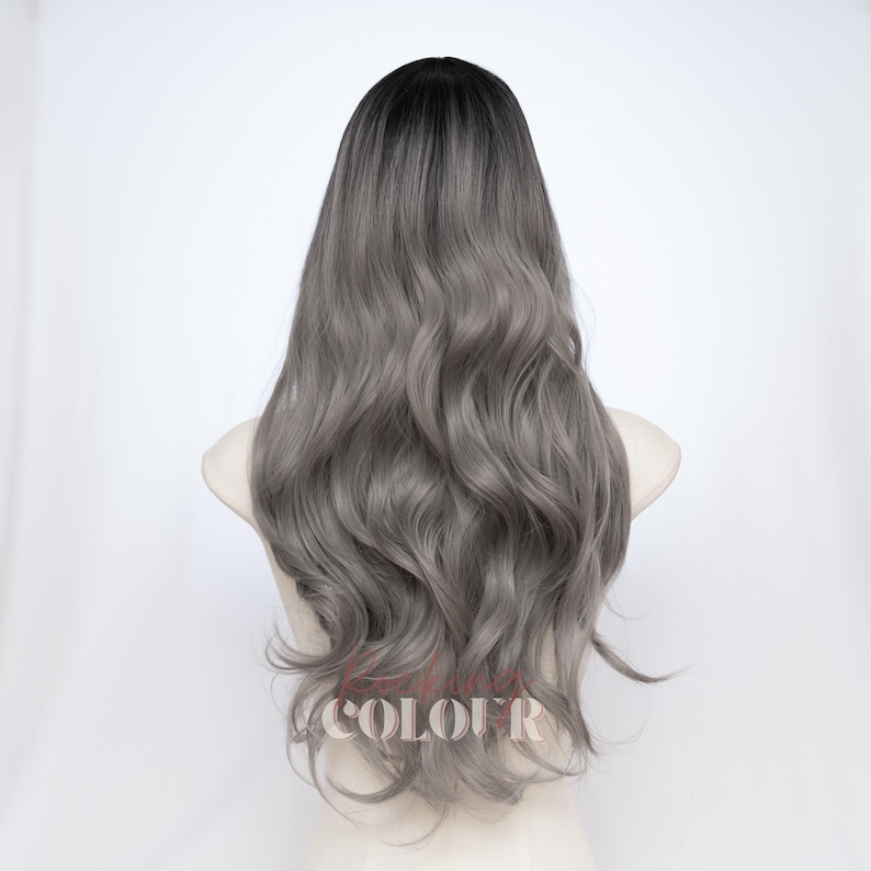 May include: A long, wavy wig featuring a dark charcoal root that transitions into a lighter ash gray tone. The hair has a soft, flowing texture with loose curls cascading down the back. The text COLOUR is visible in a light font at the bottom.