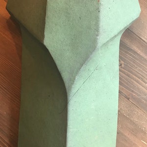 May include: A green ceramic roof tile with a unique, geometric shape. The tile has a rough, textured surface and a slightly faded appearance.