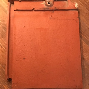 May include: A red clay roof tile with a hole in the top center. The tile has a rough texture and is slightly chipped on the edges.