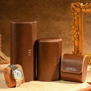 Puede incluir: Una colección de estuches de reloj de cuero marrón y un reloj de pulsera dorado. Los estuches son cilíndricos y rectangulares, con iniciales personalizadas. Un espejo con marco dorado y un martillo de madera también están en la imagen.