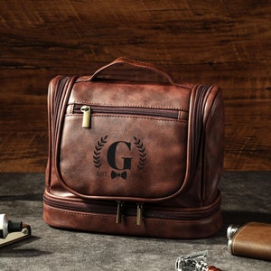 Könnte beinhalten: Eine braune Leder-Kulturtasche mit Griff, Reißverschlüssen und einem dekorativen Emblem mit dem Buchstaben "G" und einer Fliege. Ein Rasierpinsel, ein Rasierer und eine Flasche sind ebenfalls sichtbar.