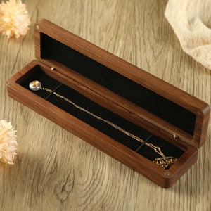 May include: An open, rectangular wooden jewelry box with a black velvet interior. A gold necklace with a pearl pendant rests inside. The box is on a light wood surface, with flowers and fabric in the background.