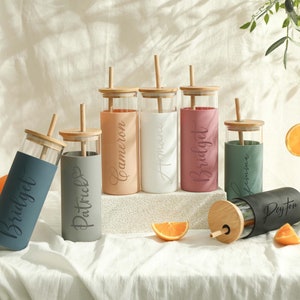 Könnte beinhalten: Sechs personalisierte Glasbecher mit Silikonschläuchen und Bambusdeckeln und -strohhalmen. Die Becher sind in verschiedenen Farben, darunter Blau, Grau, Rosa, Weiß und Grün. Jeder Becher hat einen Namen darauf, darunter Bridget, Patrick, Cameron, Andrew, Bridget und Leanne. Die Becher stehen auf einer weißen Oberfläche mit einem schwarzen Becher mit dem Namen Payton.