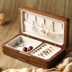 Puede incluir: Caja de joyería de madera abierta con interior claro. La caja contiene anillos, collares y pendientes. La caja es de madera oscura y tiene forma rectangular. El interior está forrado con una tela suave y blanca.