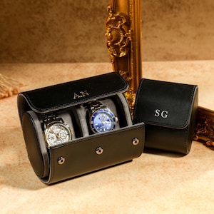 Op de afbeelding: Twee zwarte leren horloge-rollen met de initialen "A.N." en "S.G." De rol aan de linkerkant heeft twee horloges erin, een met een zilveren wijzerplaat en een met een blauwe wijzerplaat. De rol aan de rechterkant is gesloten.