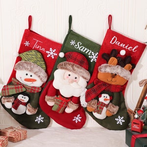 Könnte beinhalten: Drei dekorative Weihnachtsstrümpfe in Rot und Grün mit festlichen Figuren: einem Schneemann, dem Weihnachtsmann und einem Rentier. Die Strümpfe sind mit den Namen Tina, Sam und Daniel personalisiert. Schneeflocken und andere Weihnachtsdetails sind enthalten.