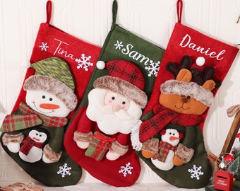 May include: Three Christmas stockings with festive designs. One red stocking features a snowman, another a Santa Claus, and the third a reindeer. Each stocking has a name embroidered on it: Tina, Sam, and Daniel. Snowflakes and other holiday details are included.