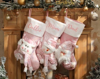 May include: Three pink Christmas stockings hang from a wooden mantel. Each stocking has a white cuff with a name embroidered in pink: Callie, Mom, and Dad. The stockings are decorated with a reindeer, snowman, and Santa Claus.