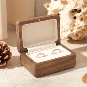 Könnte beinhalten: Eine Holz-Ringbox mit cremefarbenem Innenraum, geöffnet, um zwei goldene Ringe zu zeigen. Die Box ist aus dunklem Holz gefertigt und steht auf einer hellen Oberfläche. Eine Tannenzapfen und ein Stück Treibholz sind im Hintergrund.