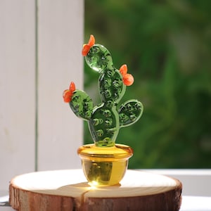 May include: A handcrafted glass cactus sculpture with a green body and orange flower accents, set in a gold-colored pot. The cactus is displayed on a wooden slice, with a blurred green background.