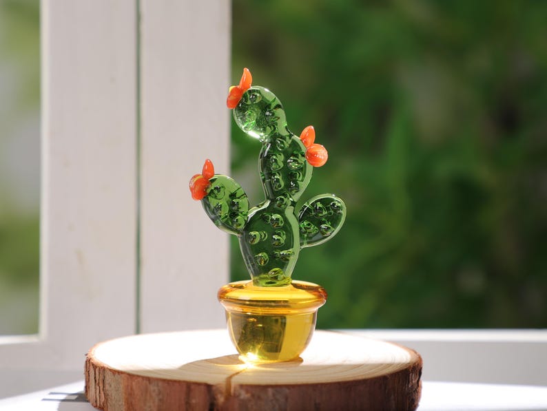 May include: A handcrafted glass cactus sculpture with a green body and orange flower accents, set in a gold-colored pot. The cactus is displayed on a wooden slice, with a blurred green background.