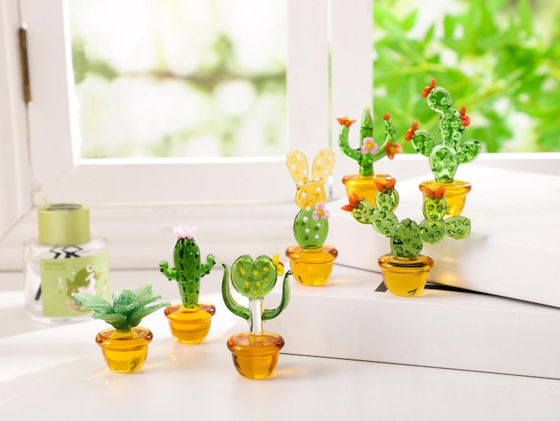 May include: A collection of miniature glass cactus sculptures in various shapes and sizes, each potted in a small yellow pot. The cacti are green with red and pink accents, and some have flower details.