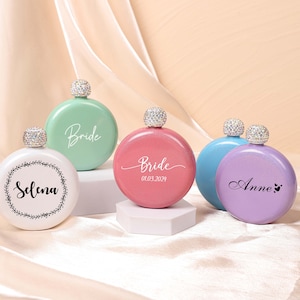 Peut inclure: Quatre flacons ronds, personnalisés, métalliques avec des bouchons ornés de bijoux. Les flacons sont de couleur blanche, verte, rose et violette. Les flacons sont étiquetés avec les noms "Selena", "Bride", "Bride 01.03.2024" et "Anne".