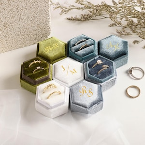 Peut inclure: Plusieurs boîtes à bagues hexagonales en velours de différentes couleurs, dont vert, bleu et blanc. Chaque boîte est brodée d'initiales en fil d'or ou blanc. Plusieurs bagues sont présentées à l'intérieur et à l'extérieur des boîtes, sur le thème du mariage.