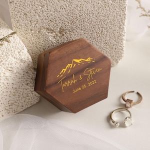 Könnte beinhalten: Eine sechseckige Holz-Ringbox mit goldener Berg-Grafik und dem Text "Tarrah & Steven June 25, 2022". Zwei Ringe mit Edelsteinen liegen neben der Box. Die Box ist braun und der Text ist goldfarben.