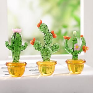 May include: Three miniature glass cactus sculptures with green bodies and orange or pink flower accents sit in yellow pots. The cacti are displayed on a white surface with a blurred green background.