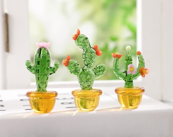 Puede incluir: Tres esculturas de cactus de vidrio en miniatura con cuerpos verdes y acentos de flores naranjas o rosas se encuentran en macetas amarillas. Los cactus se exhiben sobre una superficie blanca con un fondo verde borroso.