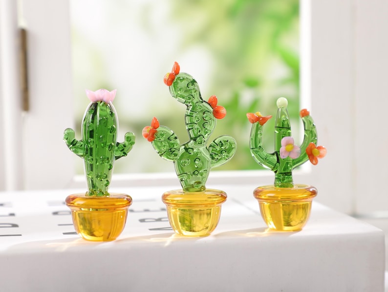 May include: Three miniature glass cactus sculptures with green bodies and orange or pink flower accents sit in yellow pots. The cacti are displayed on a white surface with a blurred green background.