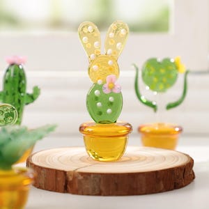 May include: A collection of glass cactus and bunny figurines. The bunny figurine is yellow with white polka dots and a green cactus body. The cactus figurines are green with pink and orange accents. They are displayed on a wooden slice and in yellow pots.
