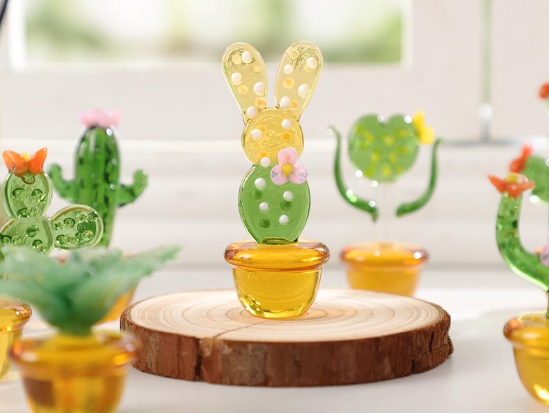May include: A collection of glass cactus and bunny figurines. The bunny figurine is yellow with white polka dots and a green cactus body. The cactus figurines are green with pink and orange accents. They are displayed on a wooden slice and in yellow pots.