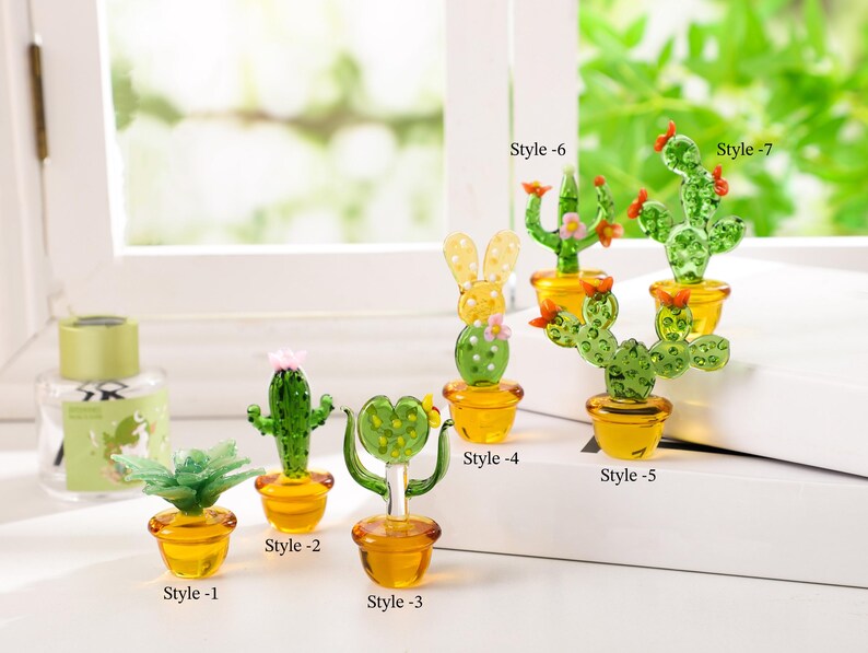 May include: A collection of miniature glass cactus sculptures in various styles, each potted in a small yellow container. The cacti are green with red and pink accents, and some have flower details. The scene is set on a white surface near a window.