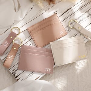 Könnte beinhalten: Eine Auswahl an personalisierten Leder-Kartenhaltern und Schlüsselanhängern in verschiedenen Rosa-, Beige- und Cremetönen. Jeder Artikel hat goldfarbene Hardware und Initialen. Die Kartenhalter haben mehrere Kartenfächer.