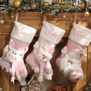 Könnte beinhalten: Drei rosa Weihnachtsstrümpfe hängen an einem hölzernen Kaminsims. Jeder Strumpf hat eine weiße Manschette mit einem rosa gestickten Namen: Saige, Jake und Kaylea. Die Strümpfe sind mit einem Rentier, einem Schneemann und dem Weihnachtsmann verziert.