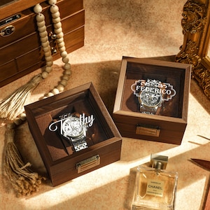 May include: Two wooden watch boxes with glass lids. The boxes are closed and feature a watch inside each. The watch boxes are on a light brown surface. The text "Federico" is engraved on one box and "Totty" is engraved on the other.