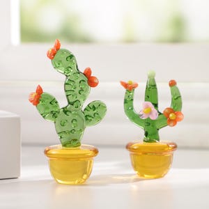 May include: Two glass cactus figurines. The cacti are green with orange and pink flower accents, set in yellow pots. The glass is transparent, allowing light to pass through. One cactus has a curved shape, while the other is upright.