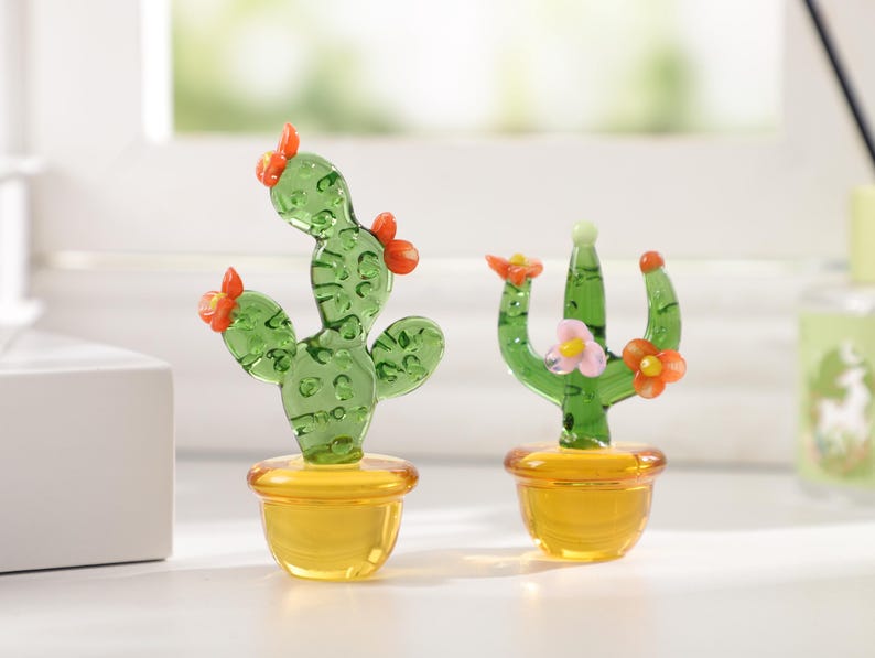 May include: Two glass cactus figurines. The cacti are green with orange and pink flower accents, set in yellow pots. The glass is transparent, allowing light to pass through. One cactus has a curved shape, while the other is upright.