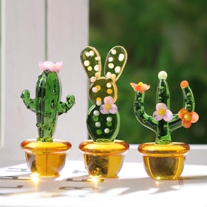 May include: Three glass cactus figurines in gold-colored pots. The cacti are green with pink and orange flower accents. The cactus on the left has a pink flower on top. The middle cactus has a bunny ear design. The right cactus has orange and pink flowers.
