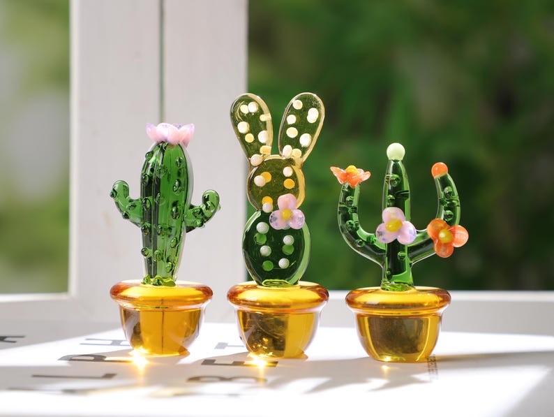 May include: Three glass cactus figurines in gold-colored pots. The cacti are green with pink and orange flower accents. The cactus on the left has a pink flower on top. The middle cactus has a bunny ear design. The right cactus has orange and pink flowers.