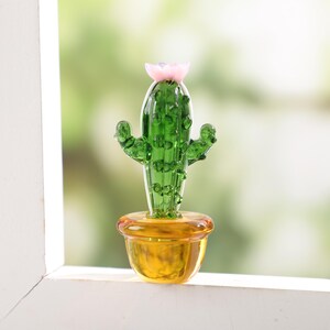 May include: A small, glass cactus sculpture with a pink flower on top. The cactus is green and sits in a yellow pot. The sculpture is displayed on a white windowsill with a blurred green background.