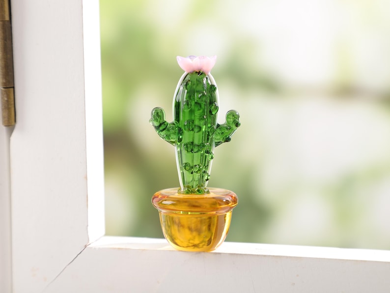 May include: A small, glass cactus sculpture with a pink flower on top. The cactus is green and sits in a yellow pot. The sculpture is displayed on a white windowsill with a blurred green background.