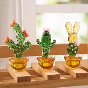 May include: Three glass cactus figurines in yellow pots sit on a wooden shelf. The cacti are green with orange and pink flower accents. The pots are a translucent yellow color, and the shelf is made of light-colored wood.