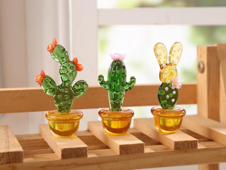 May include: Three glass cactus figurines in yellow pots sit on a wooden shelf. The cacti are green with orange and pink flower accents. The pots are a translucent yellow color, and the shelf is made of light-colored wood.